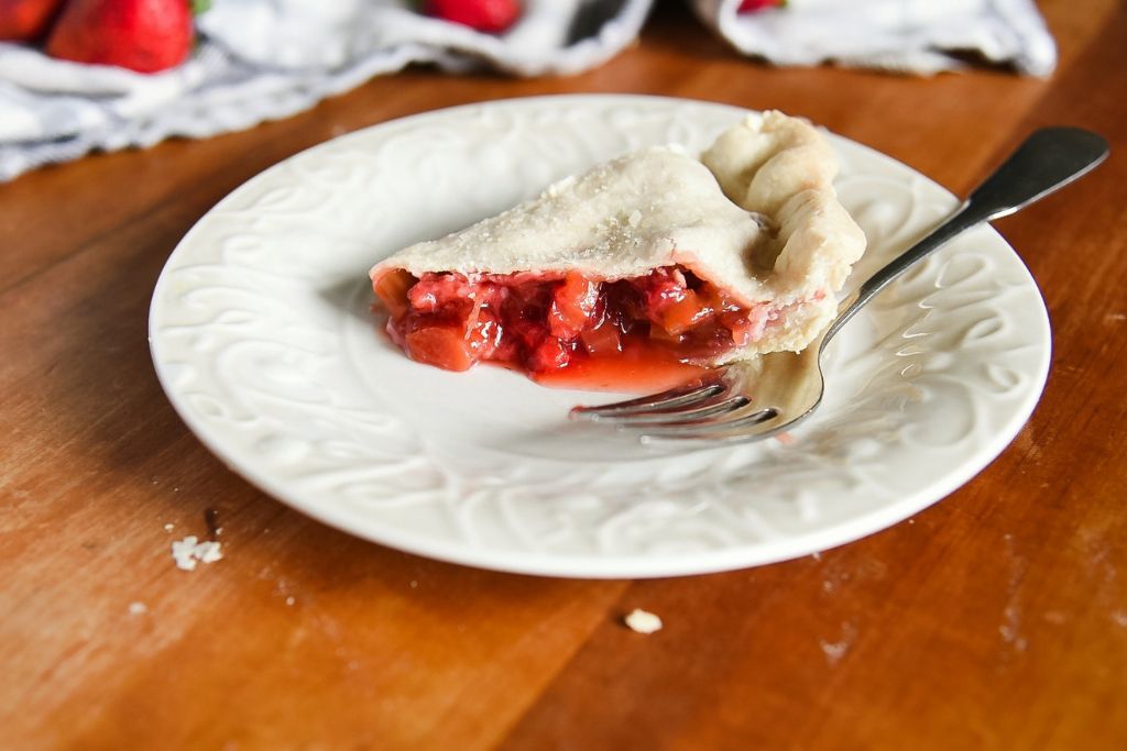 a close up of a piece of pie on a plate with a fork. There is a tea towel and some strawberries that can be seen as well.