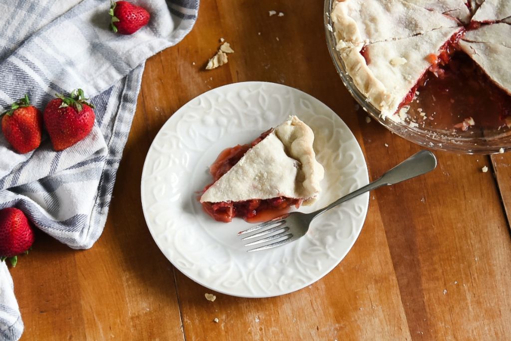a piece of pie on a plate with a fork next to it. The cut pie and a tea towel with strawberries on it in the back round