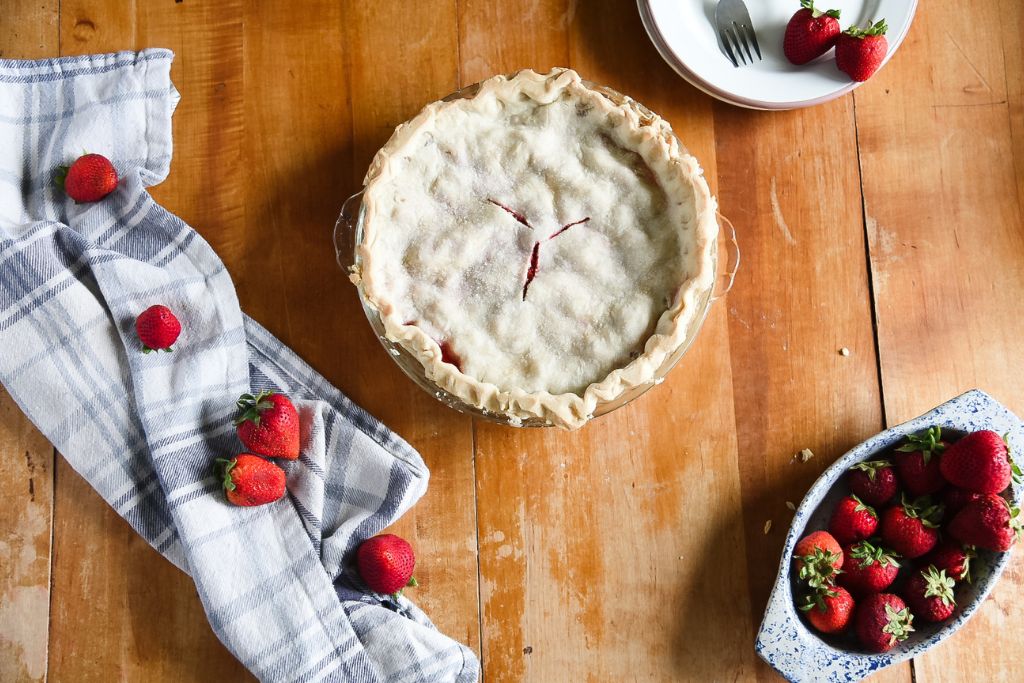 an over head shot of a pie with plates, a tea towel, and strawberries around it.
