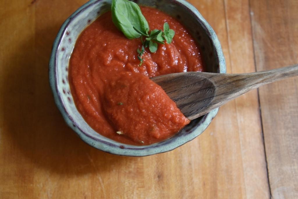 a dish of pizza sauce with herbs in it, a spoon full of sauce being taken out of the dish