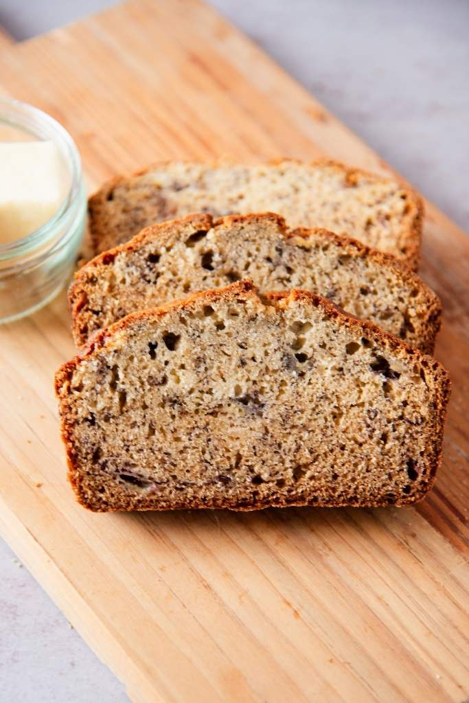 three slices on banana bread on a wooden cutting board with a dish of butter next to them