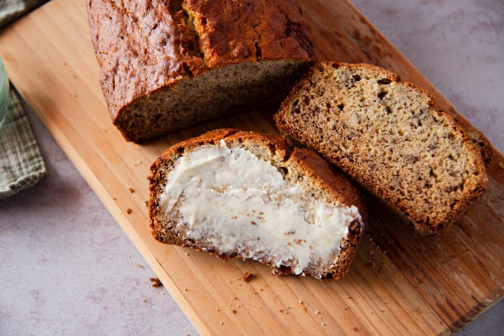 slices of banana bread one which is buttered set next to t a loaf of banana bread