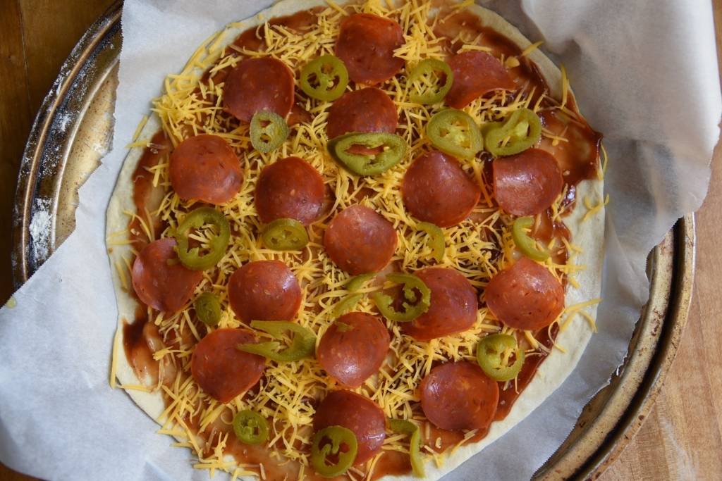 an over had shot of an uncooked pizza that has bbq sauce, cheese, pepperoni, and  Jalapeños on it
