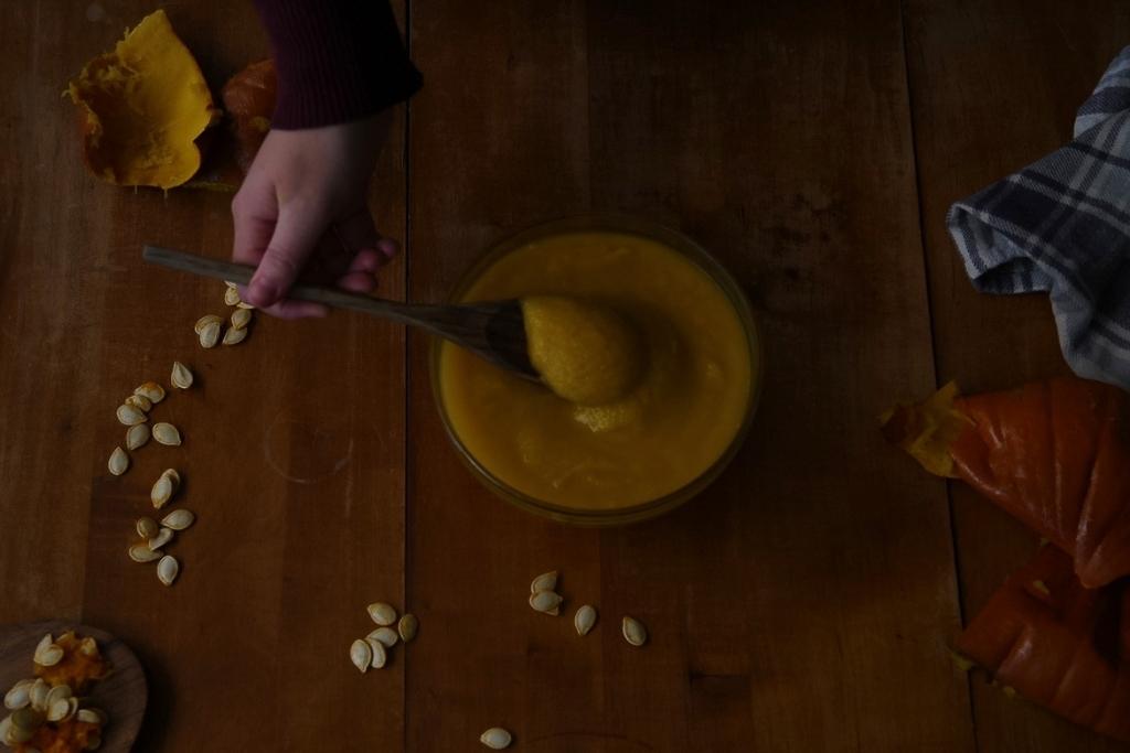 a bowl of  puree, and someone holding a spoon full of it over the bowl. There are also seeds, pumpkin peel and a towel in the back round
