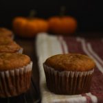 a muffin on a tea towel, there are muffins on a cooling rack next to it and some pumpkins in the back round