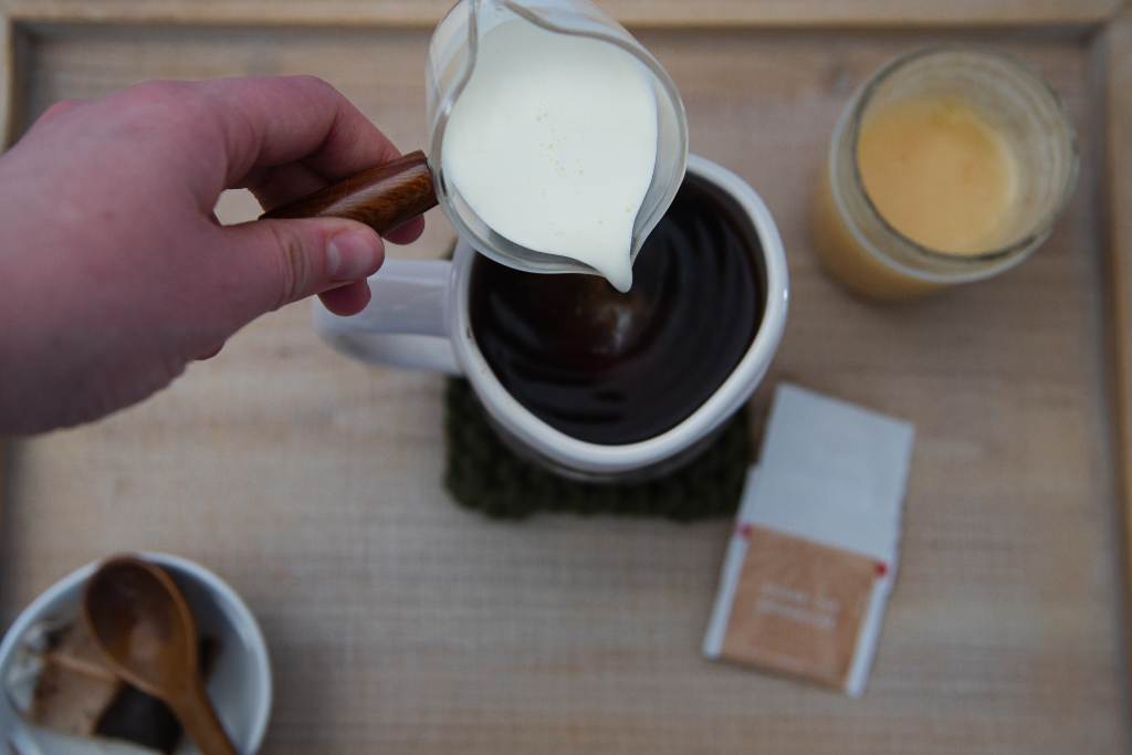 someone pouring heavy cream from a dish into a mug of black tea, there is a jar of honey, a spoon, and a dish with a tea bag on it around the mug.