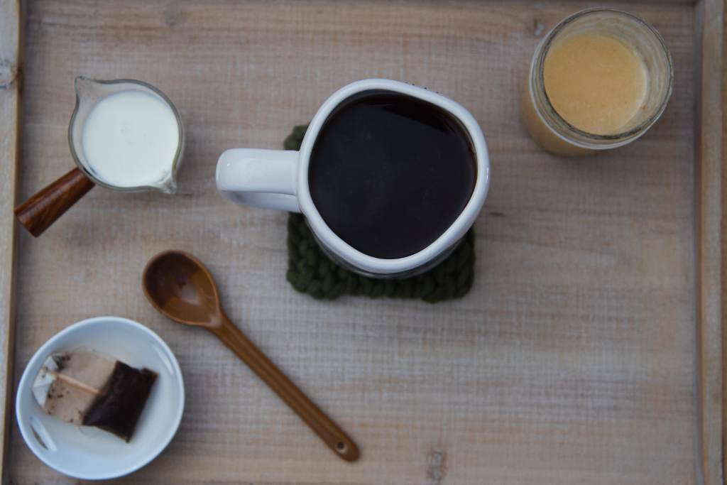 a large mug full of black tea, with a jar of honey, a dish of heavy cream, a brown spoon, a dish with a tea bag in it, around the mug