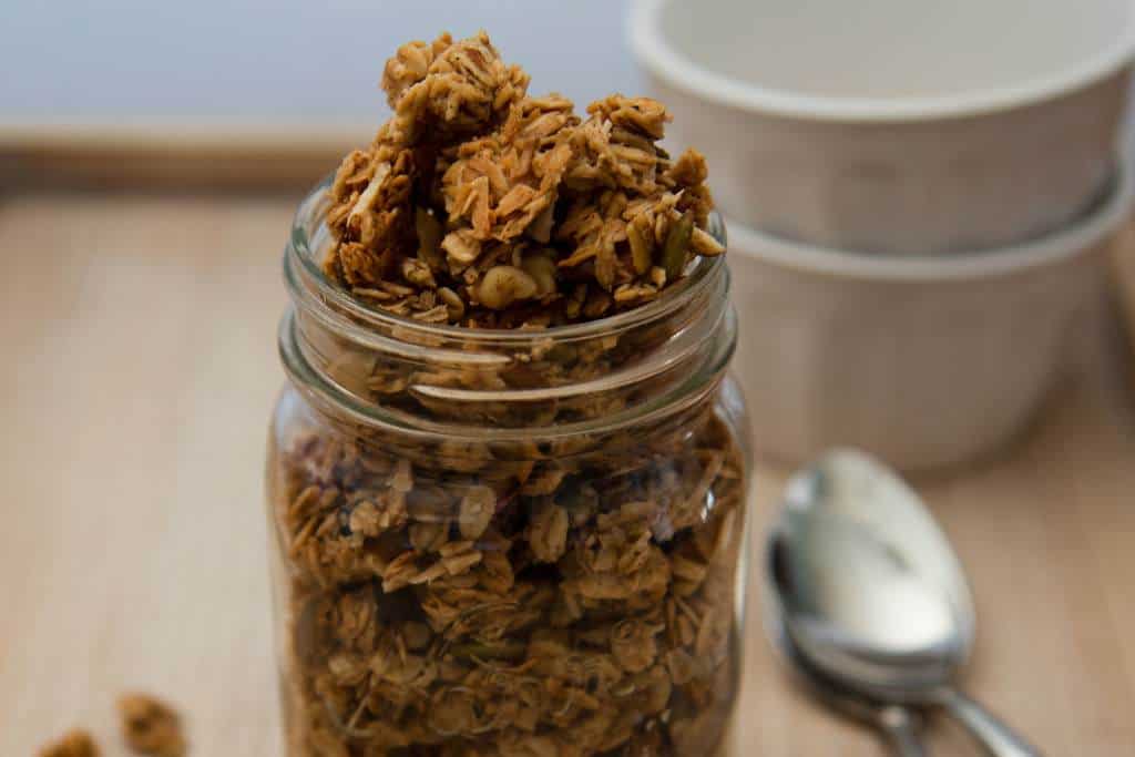 a close up of the top half of mason jar full of homemade granola, there are some dishes and spoons in the back round