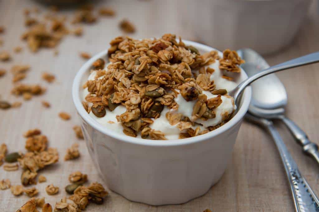 a dish full of yogurt with homemade granola sprinkled on top of it, and with the spoon in it. There is some spilled granola and some spoons in the back round