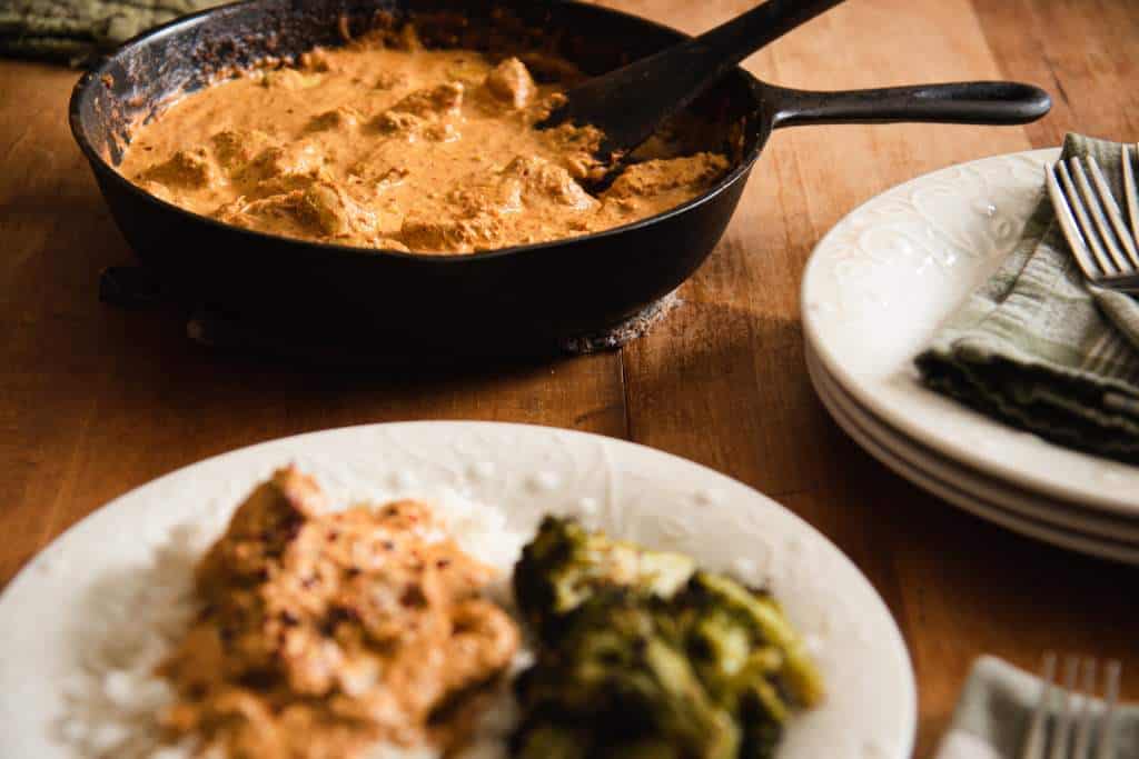 a pan of Indian butter chicken, with a plate of food, some empty plates, forks and napkins around it.