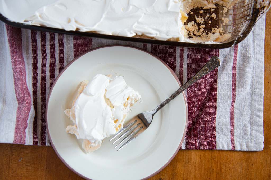 an overhead shot of a piece of banana torte on a plate with a fork, you can see the pan of torte next to the plate and it is sitting on  towel