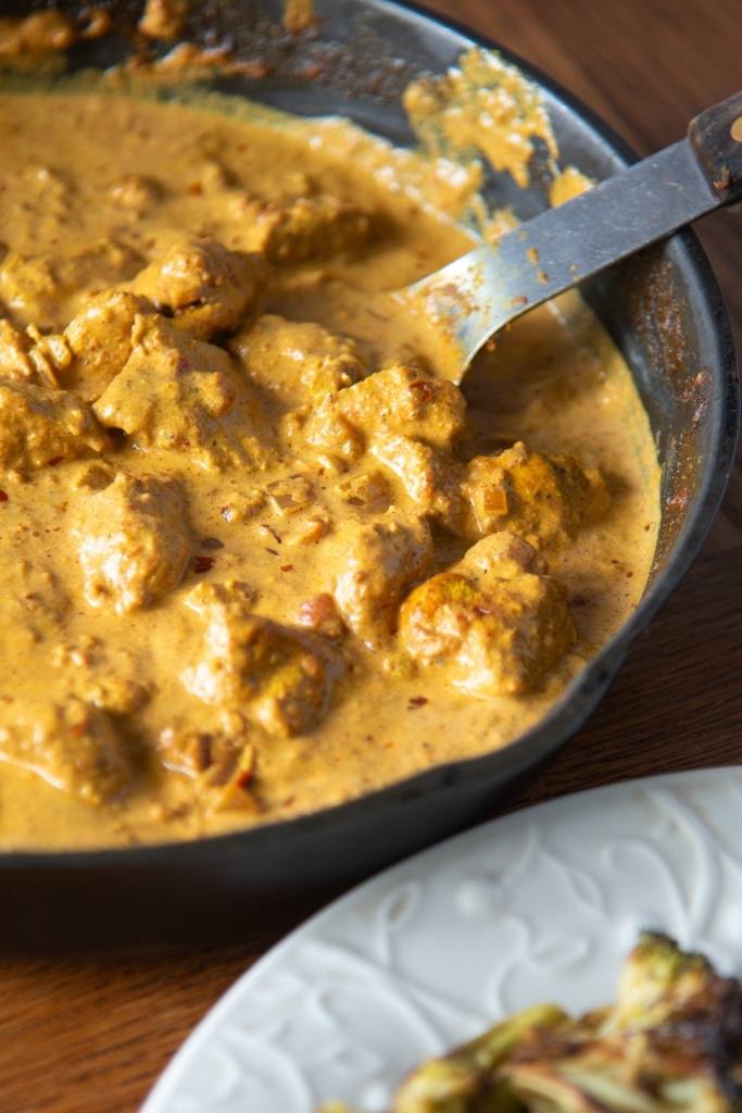 a pan of butter chicken with a spoon in it. You can also see part of a plate of butter chicken next to the pan