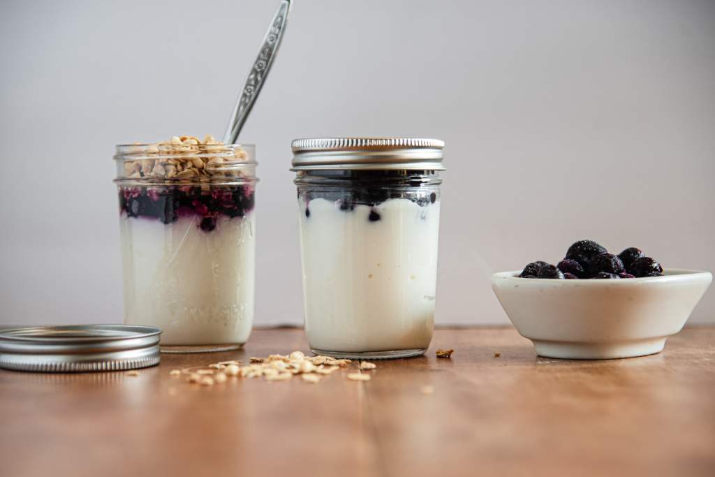 a meal prep yogurt up with yogurt blueberries, there is a lid on the jar. There is also a jar of yogurt with berries, granola and spoon in it, there is also a dish of frozen blueberries, a lid and some granola on the table