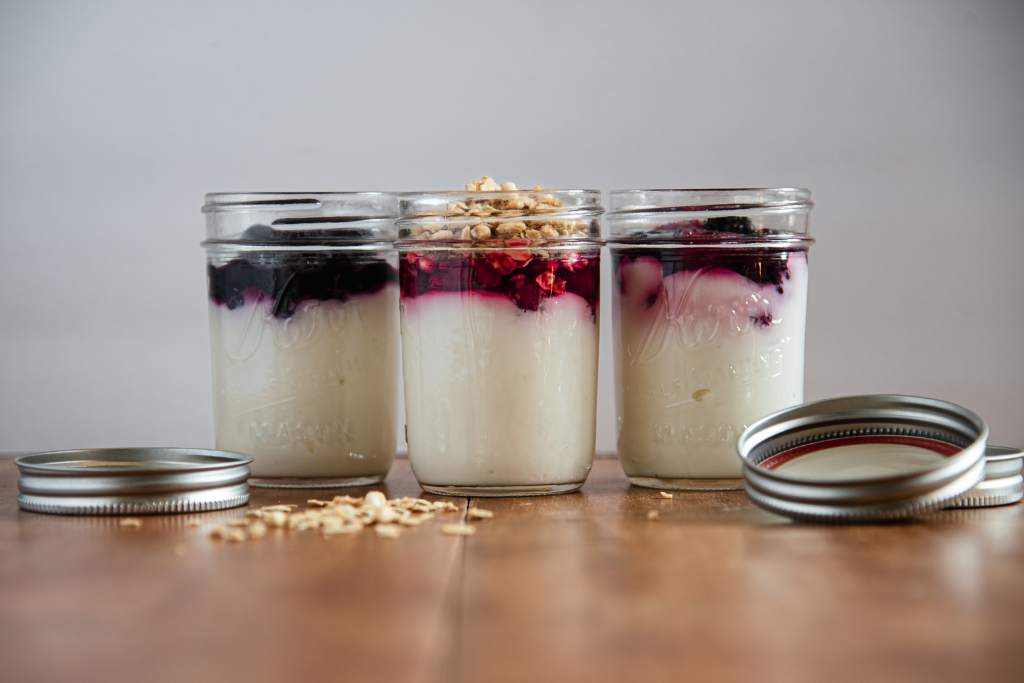 three jars of yogurt with fruit on top, there center jar has granola on it as well. There are jar lids and granola on the table in front of the jars
