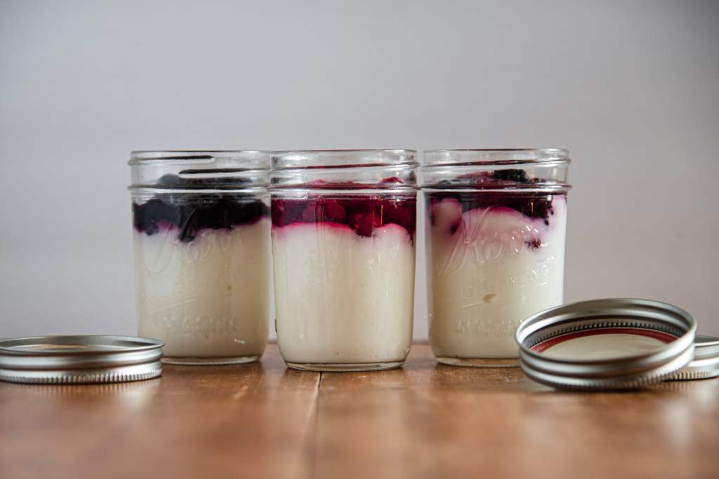 there jars on yogurt, one with raspberries, one with blueberries, and some with mixed berries. There jar lids are on the table next to the jars