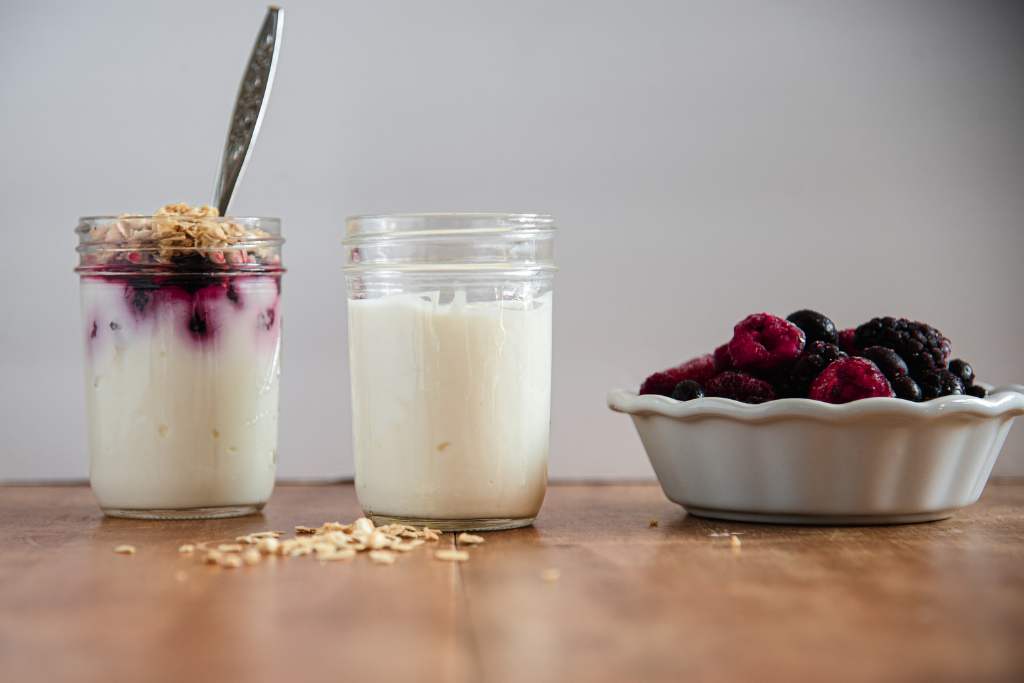 a jar of yogurt. There is a jar with yogurt, berries and a spoon in it, and a dish of mixed berries next to the jar. There is also some granola sprinkled on the table