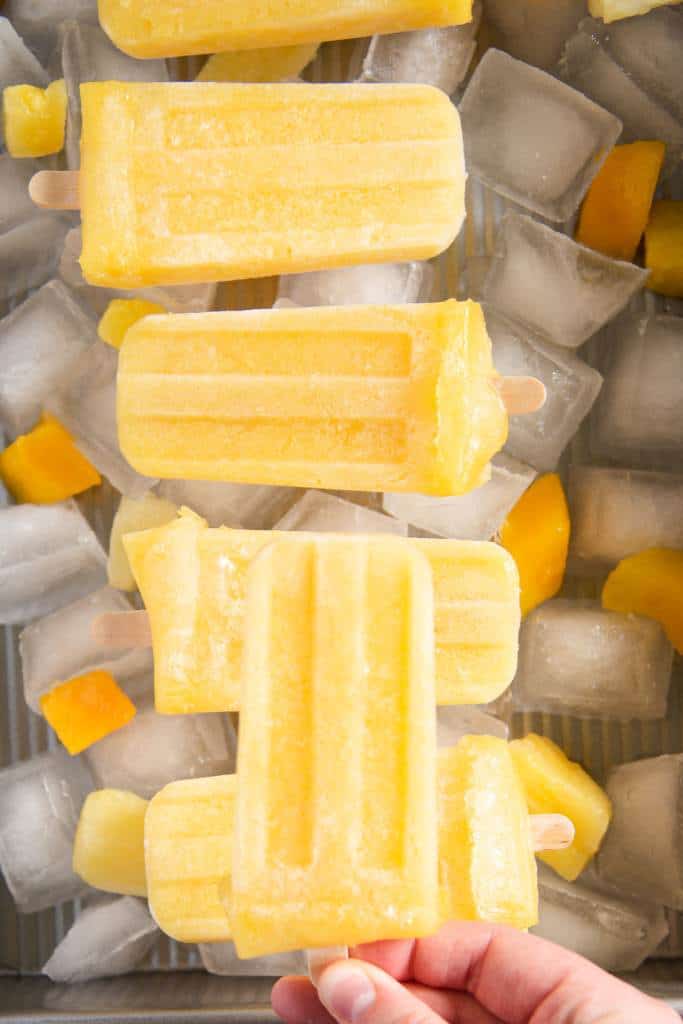 an up close of a dish with ice and fruit in it, there are five popsicles laying on the ice, and someone is holding a Popsicle over the dish 