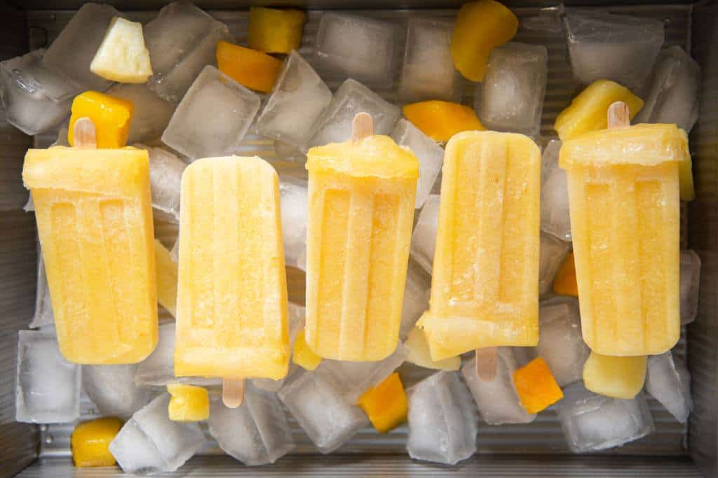 five popsicles laying on a dish of ice. there are some pineapple and mango chunks around them