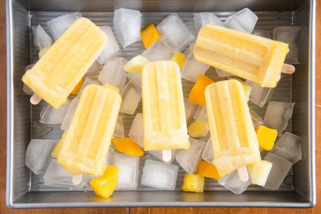 a baking dish filled with ice, and chucks of frozen fruit, there are five popsicles laying on top of the ice and fruit