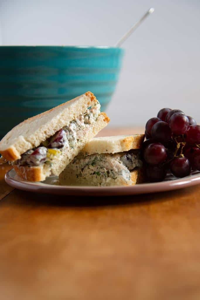 a sandwich on a plate, the sandwich has been cut in half, one half in stacked on top on the other. There are some grapes on the plate next to sandwich and there is a bowl in the background