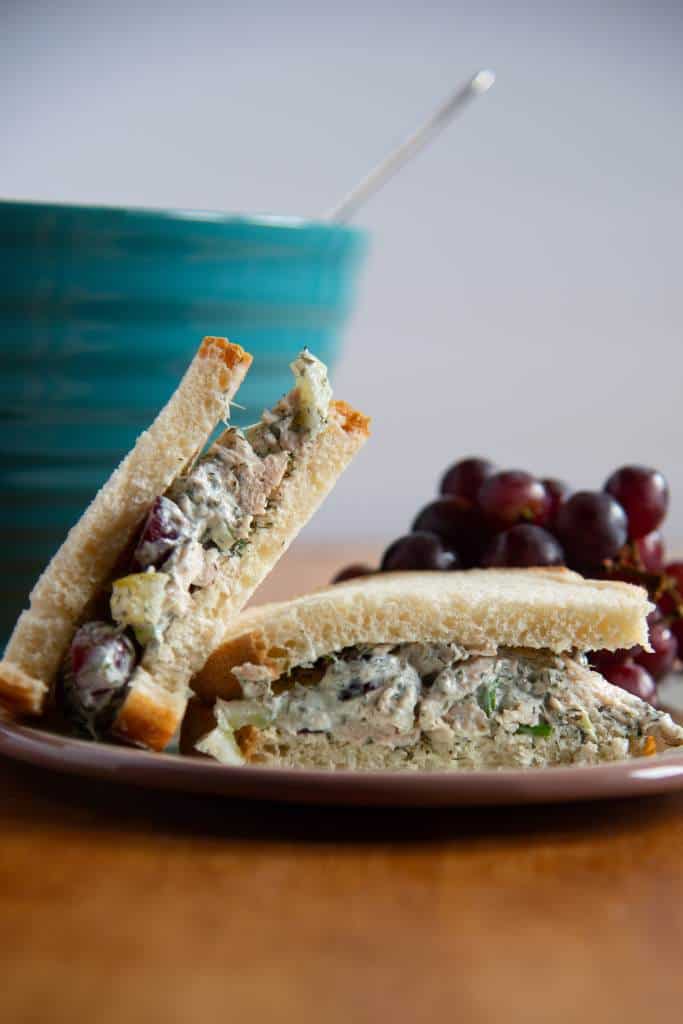 a chicken salad sandwich cut in half, one half is sitting on top of the other. There is a bunch of grapes and a bowl behind the sandwich 