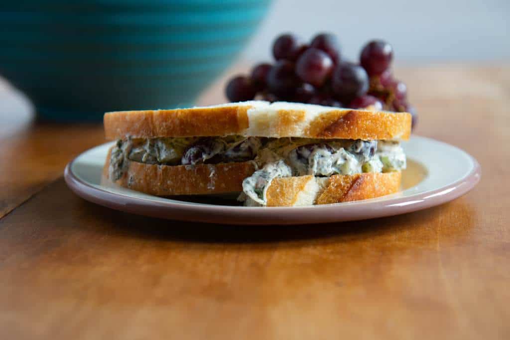 a sandwich on a plate, with a bunch of grapes and a bowl behind it.