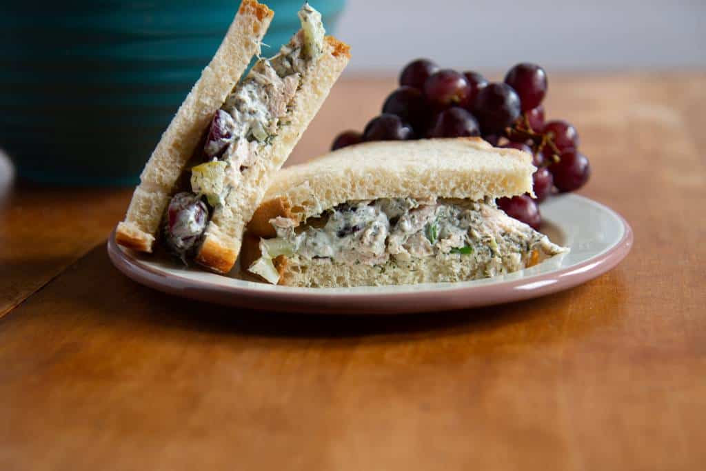 a cut chicken salad sandwich on a plate, one half is upright resting against the other have. There is a bunch of grapes on the plate as well, and a bowl in the background.