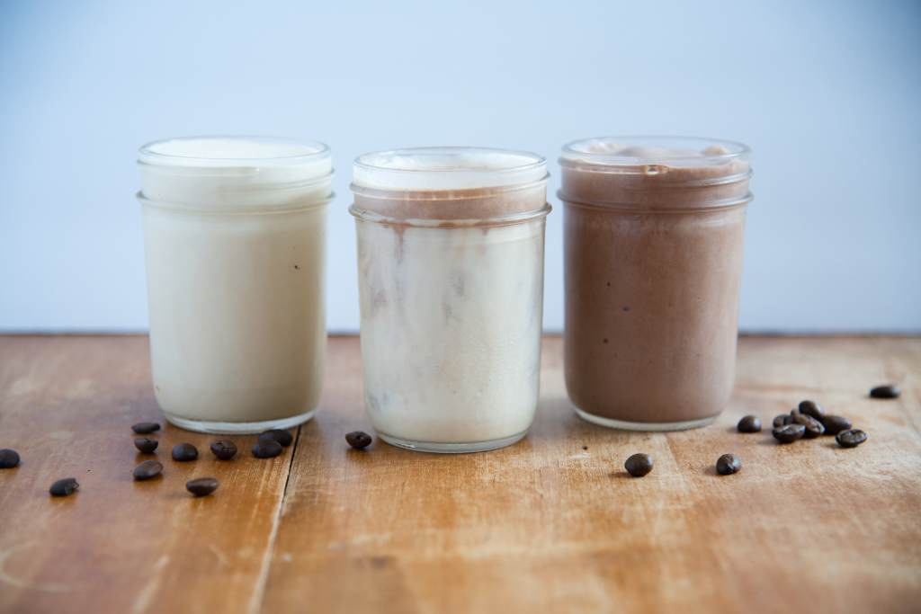 three espresso milkshakes in a life, vanilla, mixed and chocolate. There are some beans next to them on the table