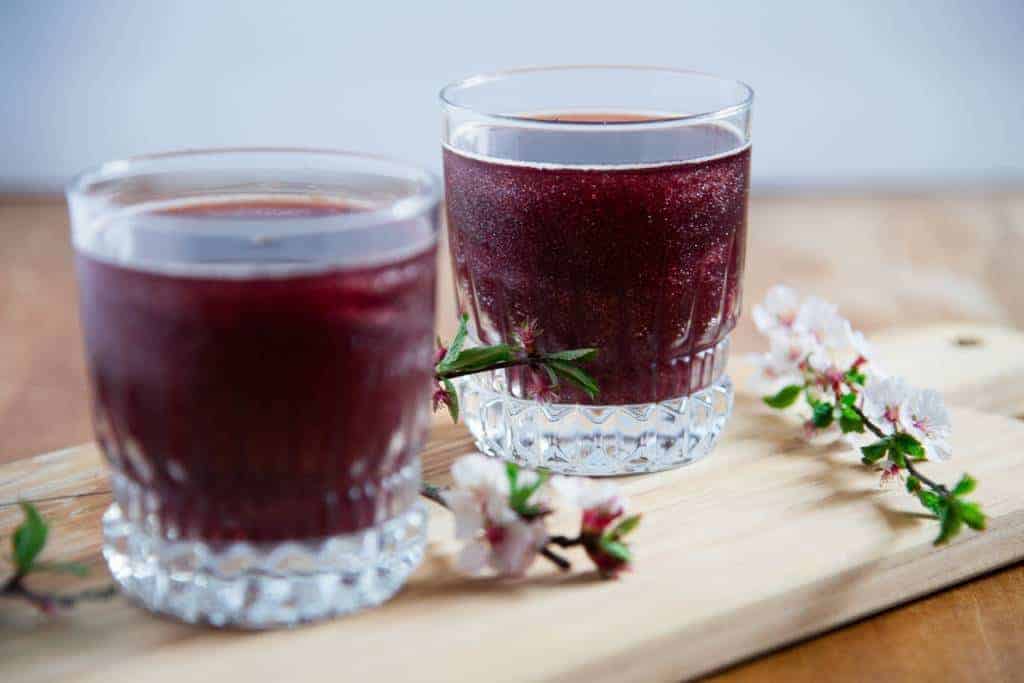 two purple rain cocktails on a board, there are some flowers around them, and the focus in on the glass in the back