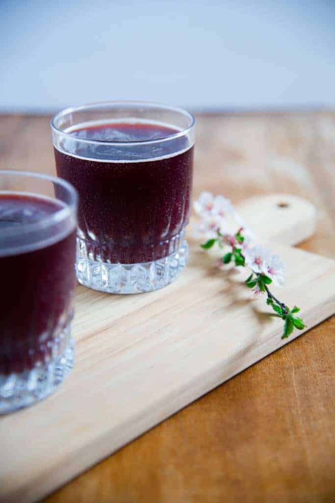 a purple rain cocktail on a board with a flowering branch next to it