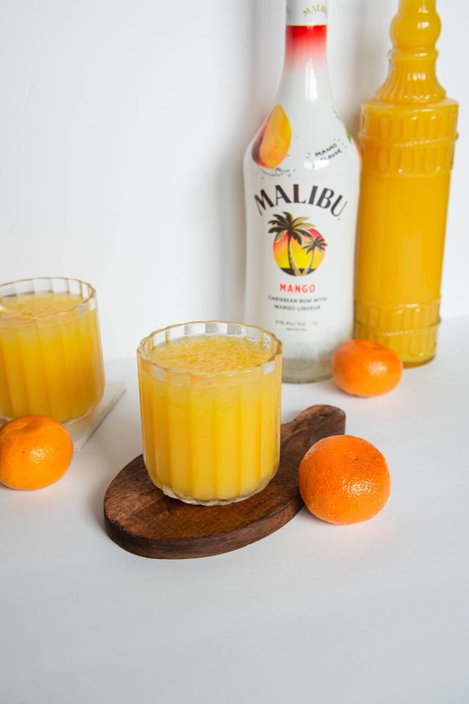 two cocktails on coasters, there are some oranges around them, and a bottle of oranges juice and a bottle of mango rum behind them 