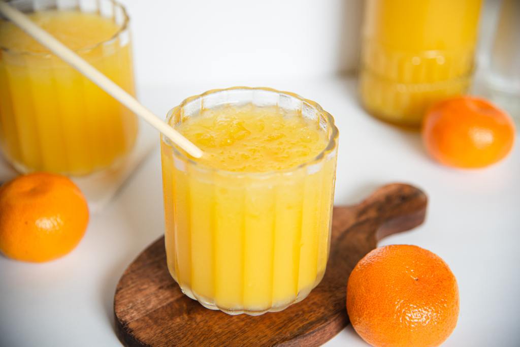 close up of a mango cocktail on a coaster, there there stir stick in the cocktail and an orange resting on the coaster, you can see another cocktail and part of a bottle in the background 