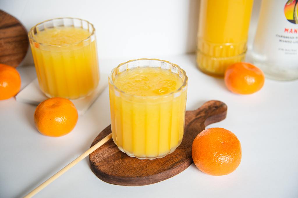 a finished cocktail on a coaster, there is another cocktail, a bottle of orange juice and bottle rum, and some oranges around it. 