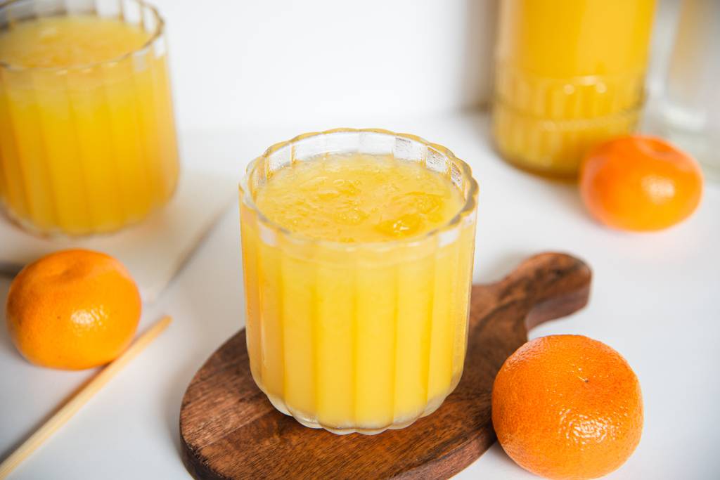 a close up picture of a mango cocktail, you can see a stair stick, some one oranges, another cocktail and part of a bottle in the background  