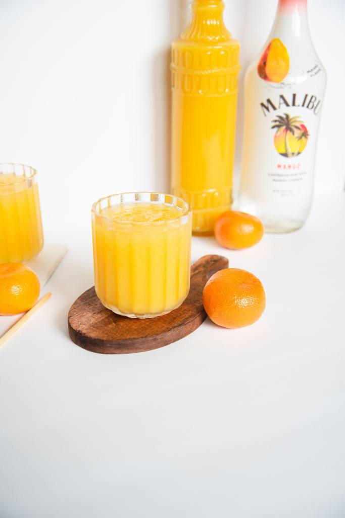 a mango cocktail on a coaster with an orange resting against it, there is bottle of orange juice and a bottle of mango rum behind and some oranges, a stir stick and another cocktail to the side of it., 