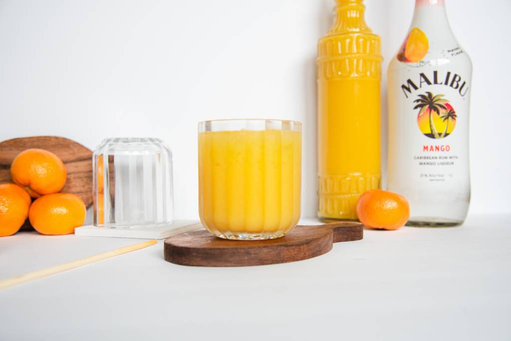 a finished cocktail on a coaster, there is a stir stick laying next to it. Behind it are some oranges, an empty glass, and a bottle of orange juice and mango rum 