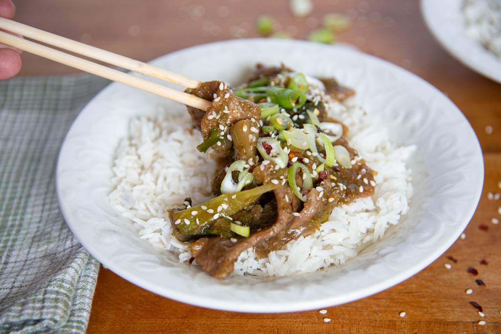 someone holding a piece of beef with chopsticks above a bowl of mongolian beef and rice