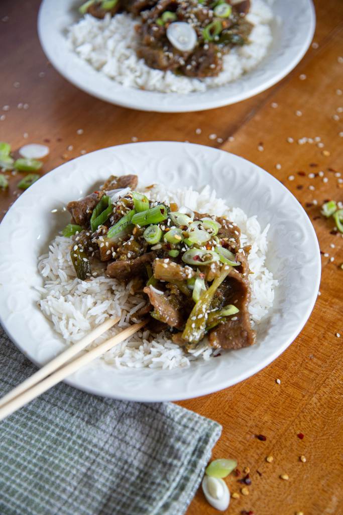 a bowl of mongolian beef and rice, there are chopsticks in the bowl, and tea towel in front of it, you ca also part of another bowl of food in the background