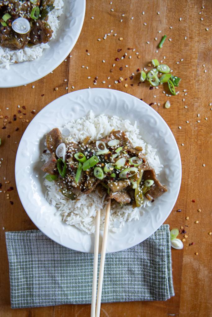 an overhead shot of a bowl of mongolian beef and rice, there is a pair of chops stick stuck in the bowl, and napkin in front of it. You can see part of another bowl of food as well