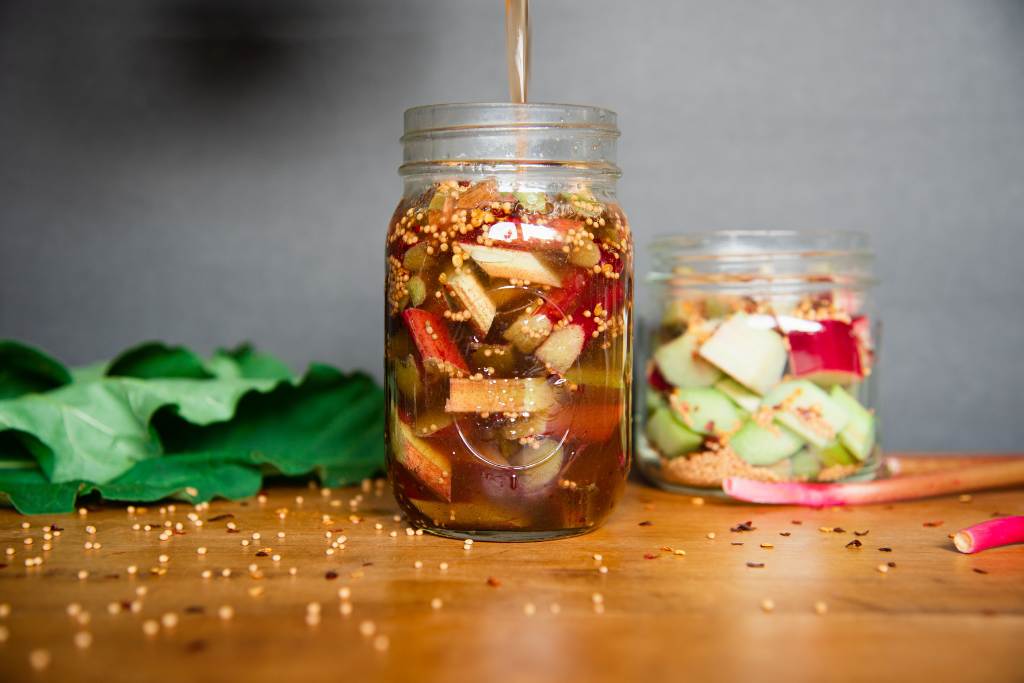 a jar of rhubarb with brine being poured in to it. There is a smaller jar behind it, as some rhubarb leaves next to it