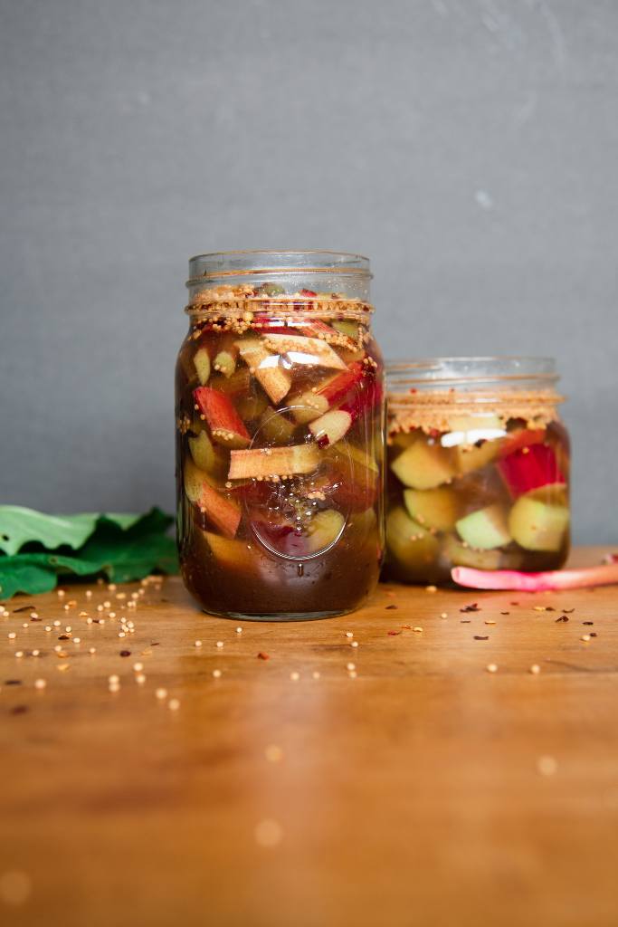 two jars of pickled rhubarb, the larger jar is in front of the smaller one. There are some stalks, leaves, and spices around them