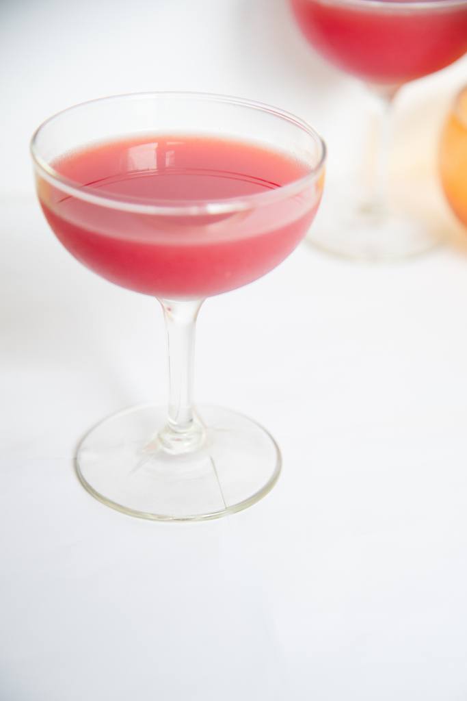 a slightly overhead shot of a finished pink cocktail, you are see another cocktail and dish behind it.