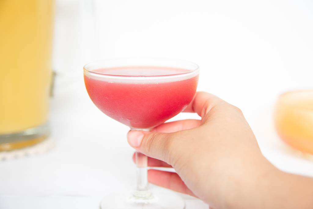 a close up of someone holding a Rosie's Cocktail, you can see part of a dish a pitcher of orange juice in the background.