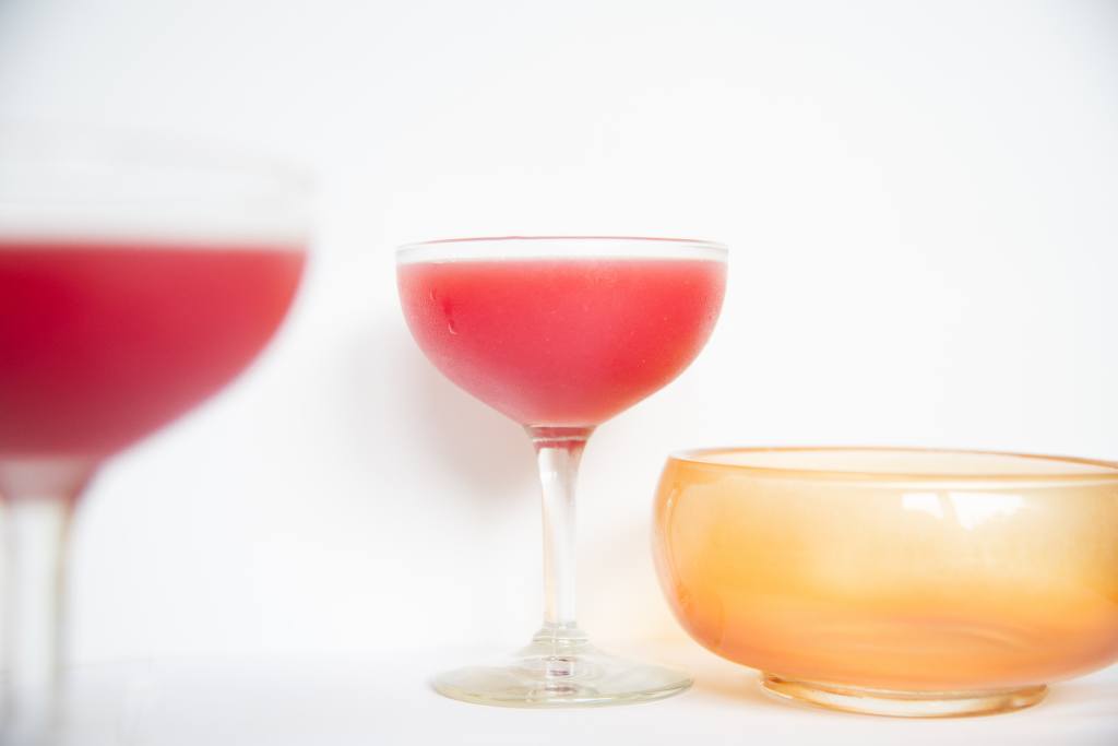 a pink cocktail, with a drop of water running down the glass. There is a dish next to it, and blurry cocktail to the side of it.