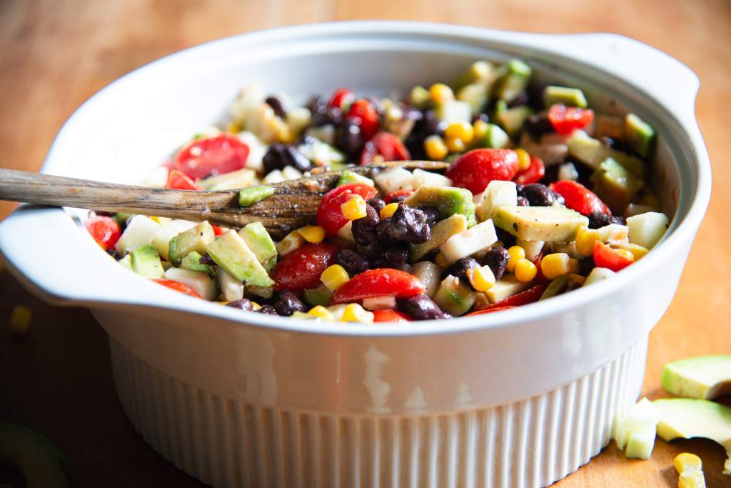 a bowl full of avocado salad with a wooden spoon in it. There are some avocado slices and corn kernels next to the bowl