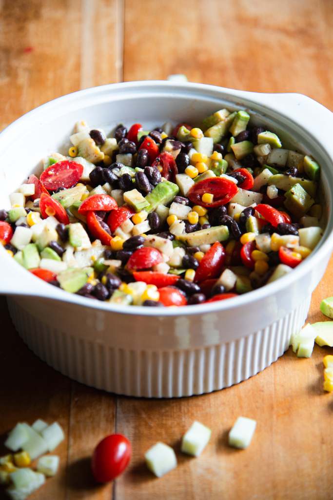 a white bowl of salad, you can part of an avocado and some corn next to it.