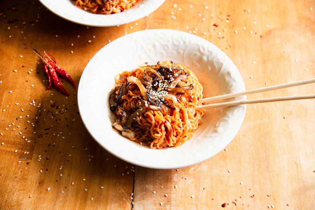 an overhead shot of a bowl of gochujang noodles with chopsticks stuck in them, there are onions and sesame seeds on top of the noodles, and some dried peppers and seasonings on the table around the bowl.