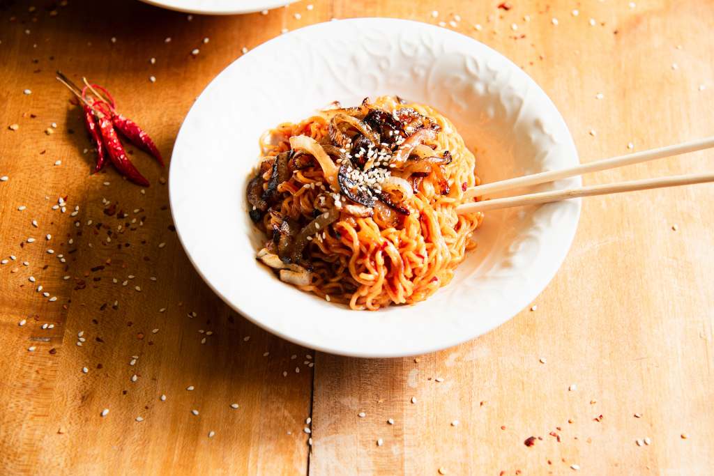 a white bowl of noodles topped with onions and sesame seeds, there are chopsticks stuck in the noodles and some dried peppers  next to the bowl