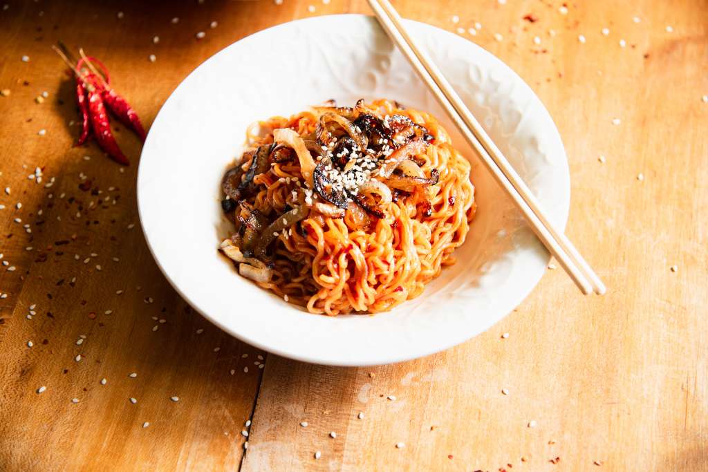 a close up of a bowl noodles topped with onions and sesame seeds, there is a pair of  chopsticks resting on the edge of the bowl, and some dried peppers and seasonings around the bowl