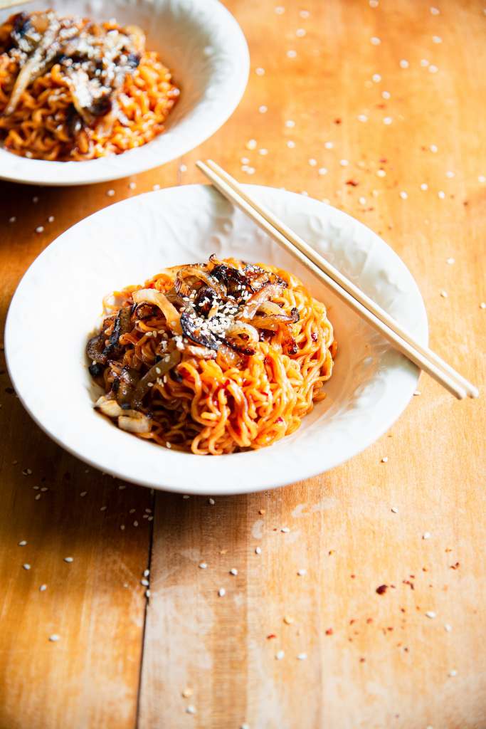 a bowl of spicy gochujang noodles with onions and sesame seeds on it. There is a set of chopsticks on the edge of the bowl, and you can see another bowl of food in the background 